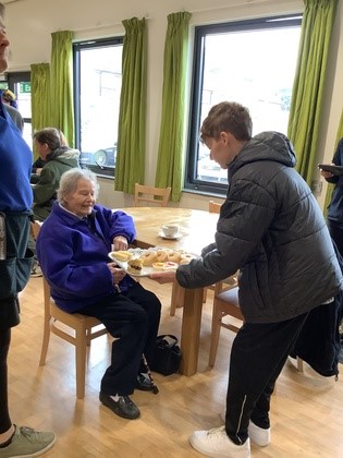 Bletchley Macmillan Coffee Morning Student Handing Cake To Visitor